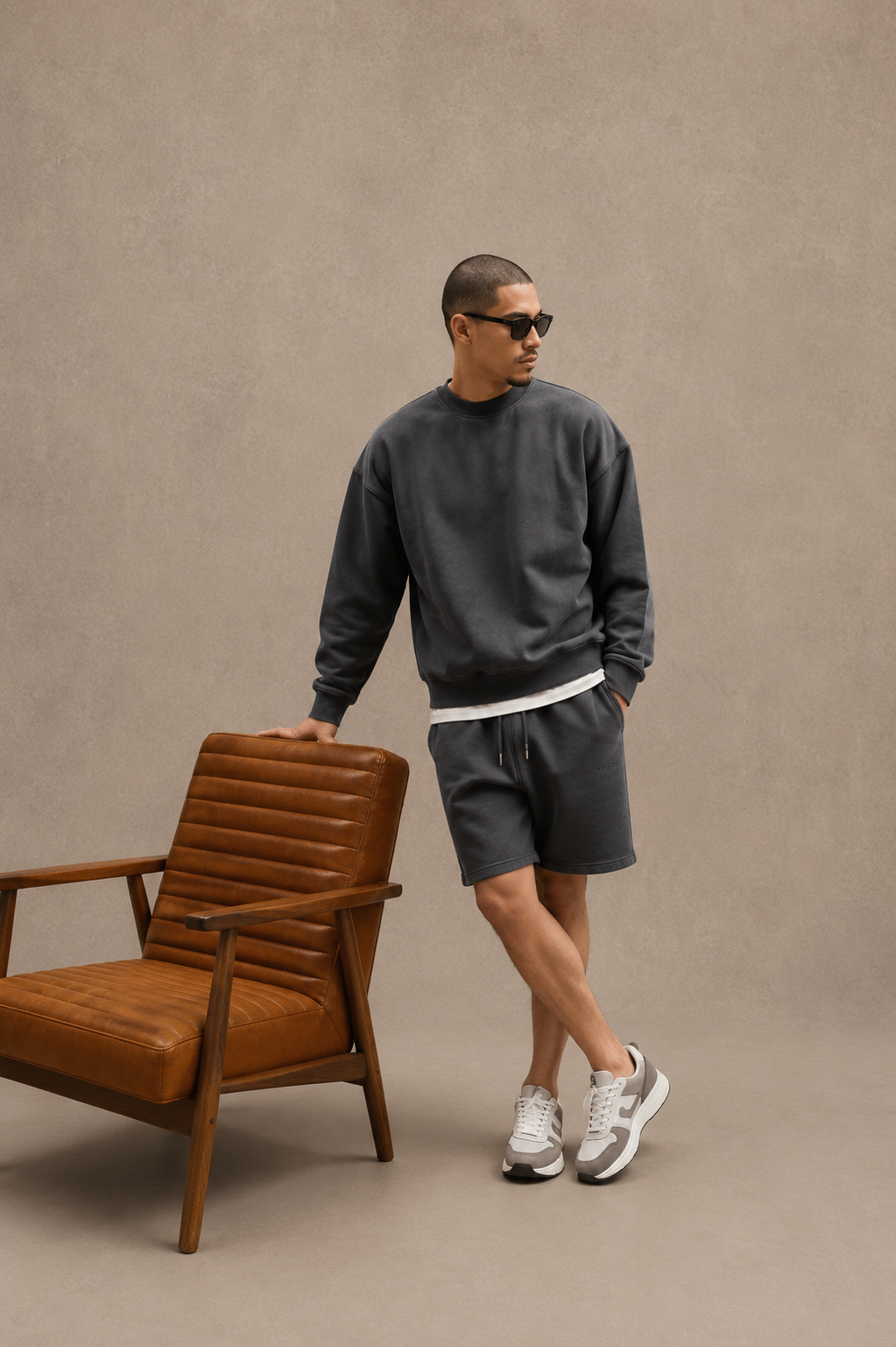 Man in dark gray sweatshirt and shorts standing next to a brown chair against a beige background