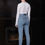 Woman wearing a white long-sleeve top and blue jeans standing in front of a dark wall.
