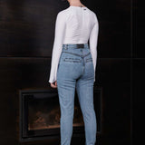Woman wearing a white long-sleeve top and blue jeans standing in front of a dark wall.