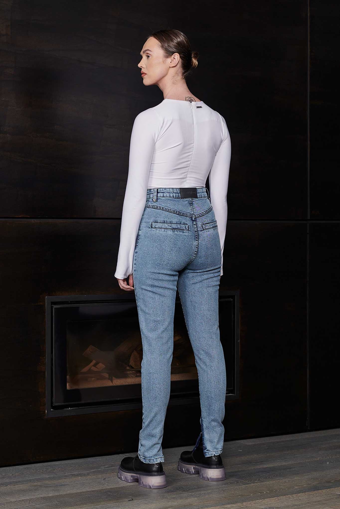 Woman wearing a white long-sleeve top and blue jeans standing in front of a dark wall.