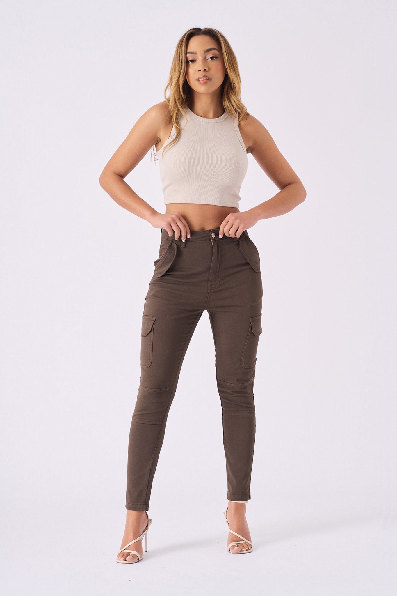 A woman posing in high waisted brown skinny cargo jeans with a beige top and clear heels.