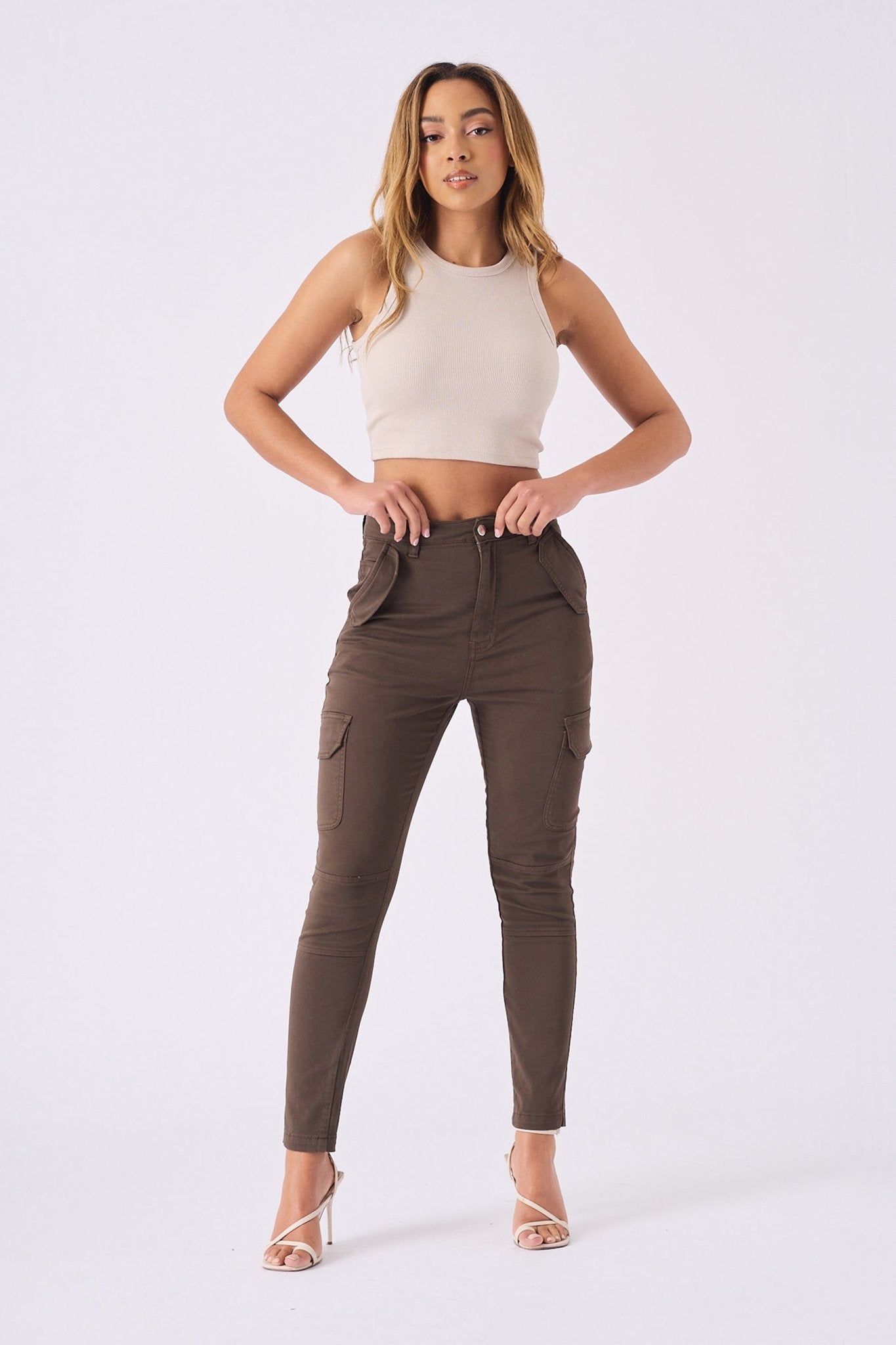 A woman posing in high waisted brown skinny cargo jeans with a beige top and clear heels.
