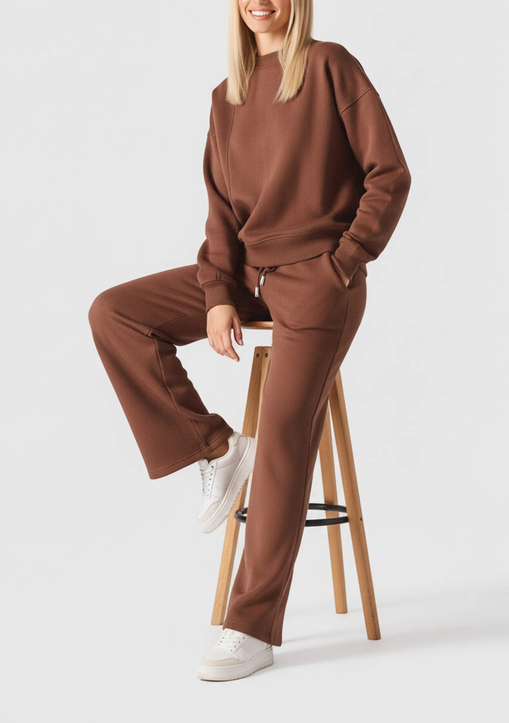 A model sitting on a stool wearing a brown crew neck jumper and wide leg trousers