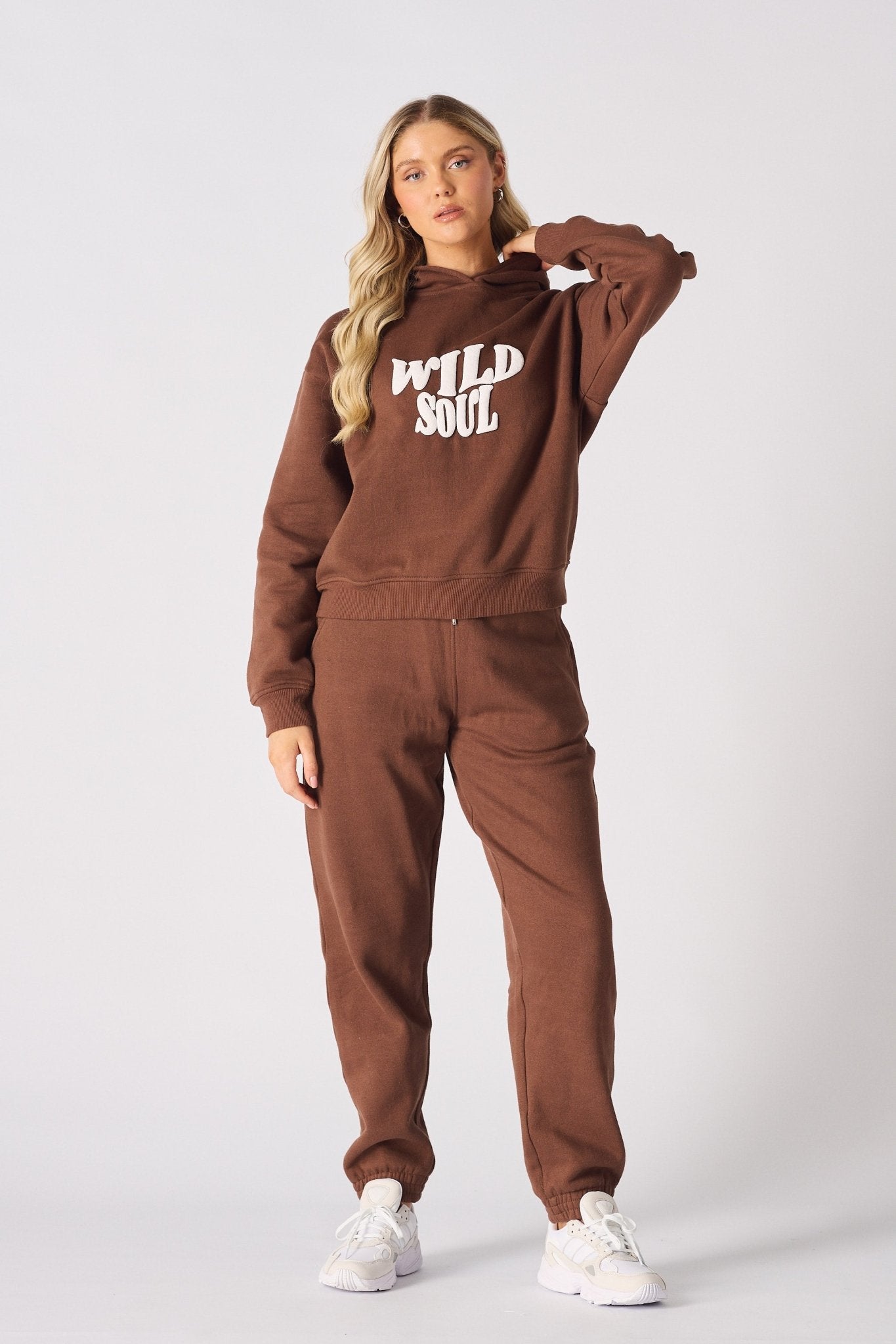 A woman wearing a brown graphic hoodie with matching joggers and white trainers