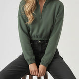 A blonde woman sitting on a stool wearing a dark green half zip sweatshirt and black jeans