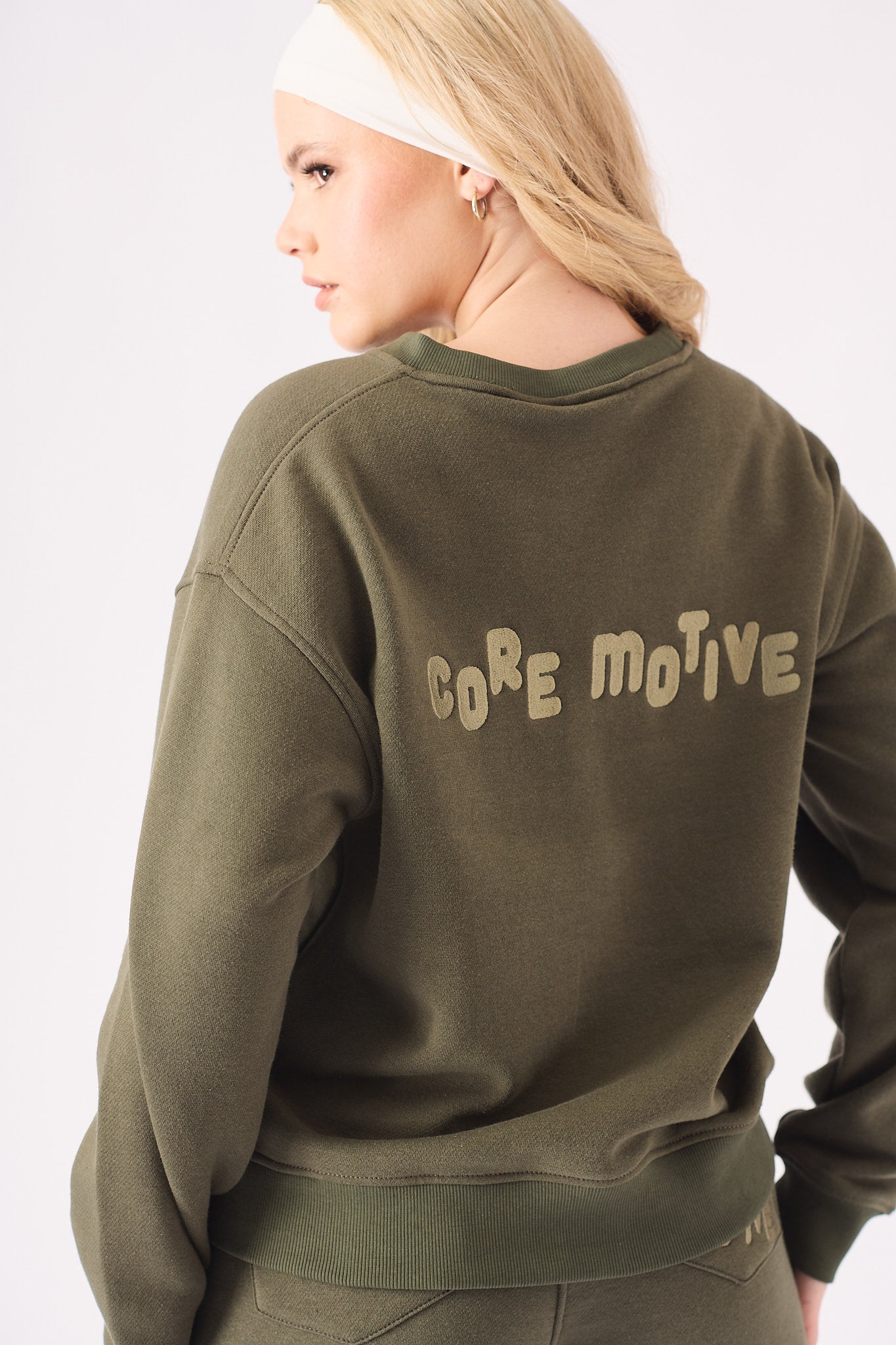 Rear view of a woman wearing a khaki crew neck jumper showing the bold core motive branding