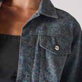 Extreme close up of speckle print denim jacket collar and chest pocket with Herman&Co branded button - showcasing mottled blue-grey fabric 