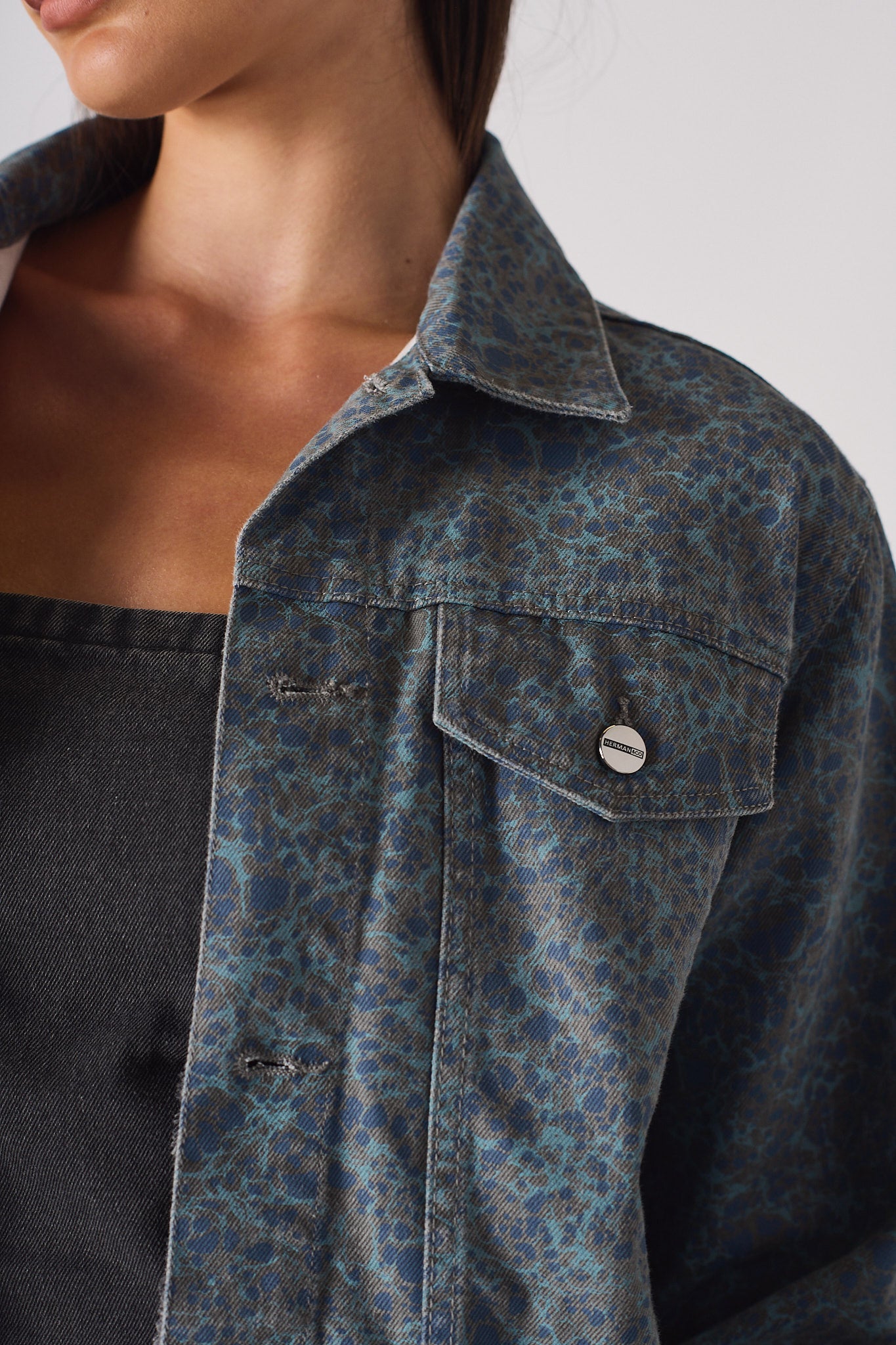 Extreme close up of speckle print denim jacket collar and chest pocket with Herman&Co branded button - showcasing mottled blue-grey fabric 