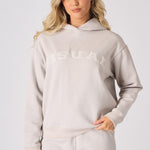 A front view of a woman wearing a stone coloured graphic hoodie with a towelling patch