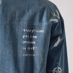 Back close up of mid wash tattoo print denim jacket showing “Everything you can imagine is real.” Pablo Picasso quote embroidery along with swallow, feather, and compass motifs on sleeve
