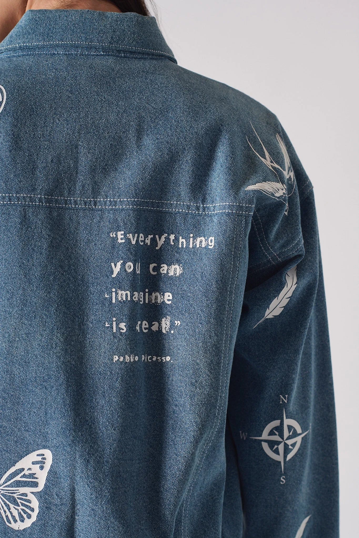 Back close up of mid wash tattoo print denim jacket showing “Everything you can imagine is real.” Pablo Picasso quote embroidery along with swallow, feather, and compass motifs on sleeve
