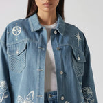 Upper body close up of mid wash tattoo print denim jacket showing collar, placket, chest pockets, and embroidered motifs including compass, star, rose, and butterfly over white tee