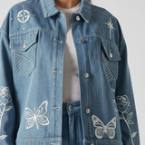 Upper body close up of mid wash tattoo print denim jacket showing collar, placket, chest pockets, and embroidered motifs including compass, star, rose, and butterfly over white tee