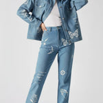 Front pose of mid wash tattoo print denim jacket with hand behind neck showing sleeve embroidery - women’s oversized set with celestial symbols and floral details on cargo pants
