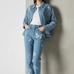 Front view of mid wash tattoo print denim jacket worn open over white tee - women’s oversized embroidered set with hands in pockets and concrete wall backdrop