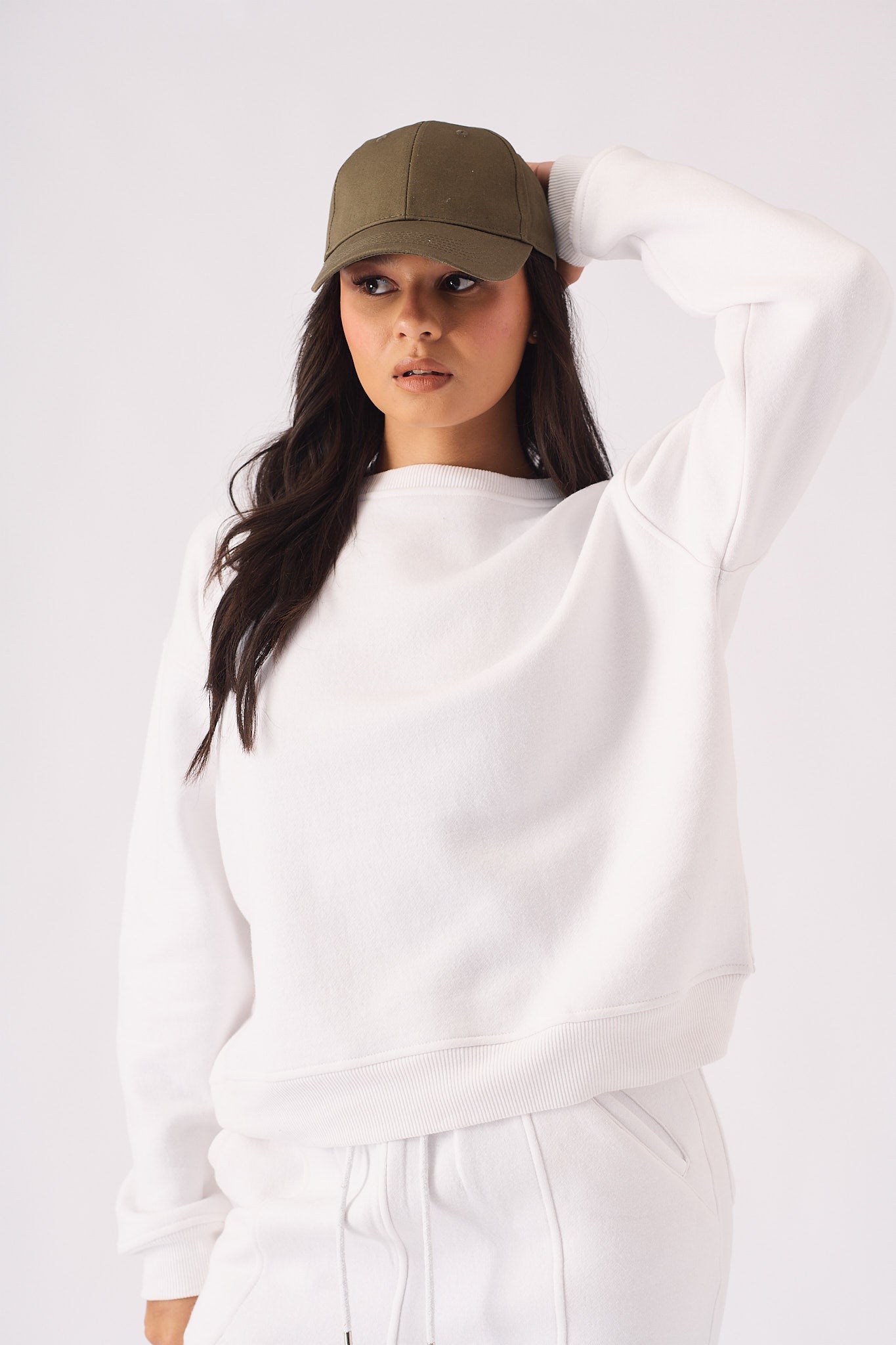 A woman posing in a white crew neck jumper while holding her cap for a sporty style