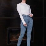 Woman wearing a white long-sleeve top and blue jeans standing in front of a dark wall.