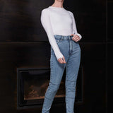 Woman wearing a white long-sleeve top and blue jeans standing in front of a dark wall.