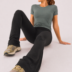 A woman seated on the floor wearing womens-black-kick-flare-leggings showing the flare detail and high waist fit, styled with a green crop tee