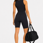 Back view of a woman wearing a black racer neck crop top and matching shorts, holding a gym bag.