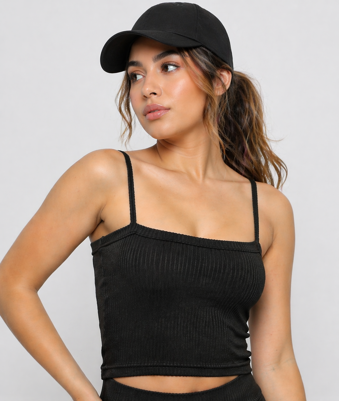 A woman wearing womens-black-ribbed-crop-top with thin cami straps and a black cap, front view