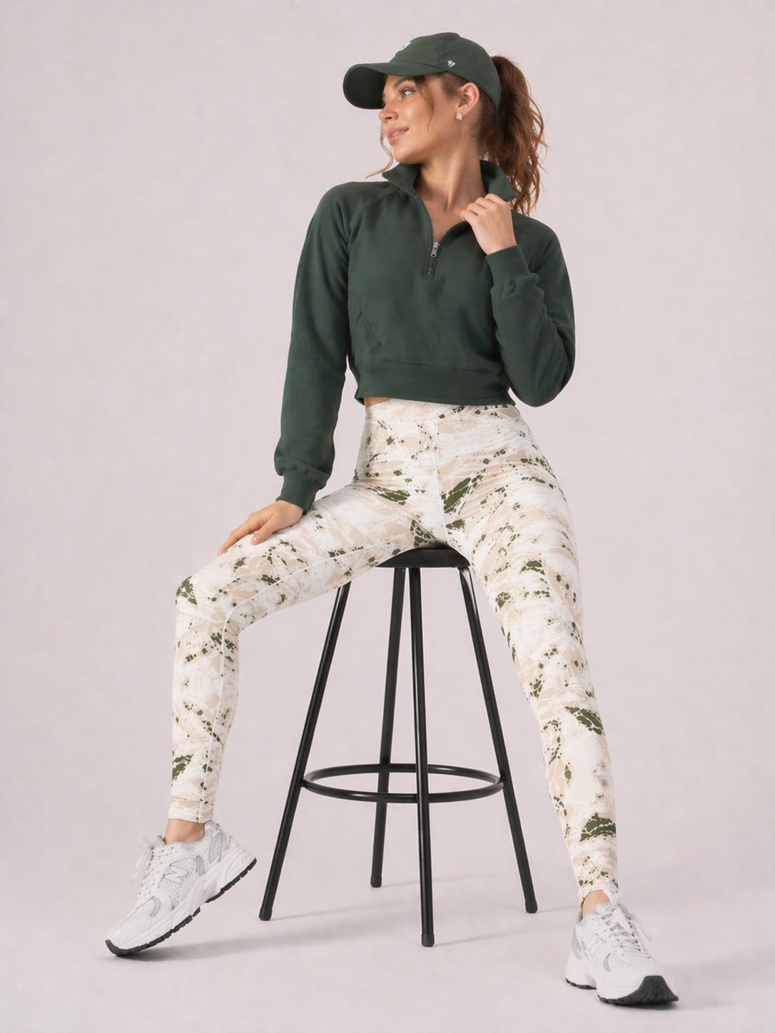 A woman seated on a stool wearing womens-green-stone-print-high-waist-leggings styled with a green half zip crop and white trainers
