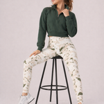 A woman seated on a stool wearing womens-green-stone-print-high-waist-leggings styled with a green half zip crop and white trainers