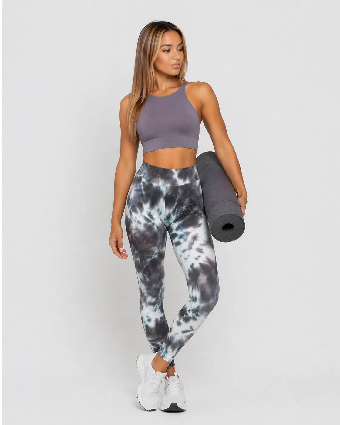 A woman wearing womens-grey-tie-dye-high-waist-leggings holding a yoga mat, front view styled with a purple crop top and white trainers