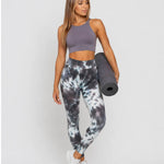 A woman wearing womens-grey-tie-dye-high-waist-leggings holding a yoga mat, front view styled with a purple crop top and white trainers