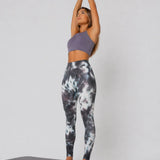 A woman in womens-grey-tie-dye-high-waist-leggings standing on a yoga mat with arms raised above head, side view showing full length fit