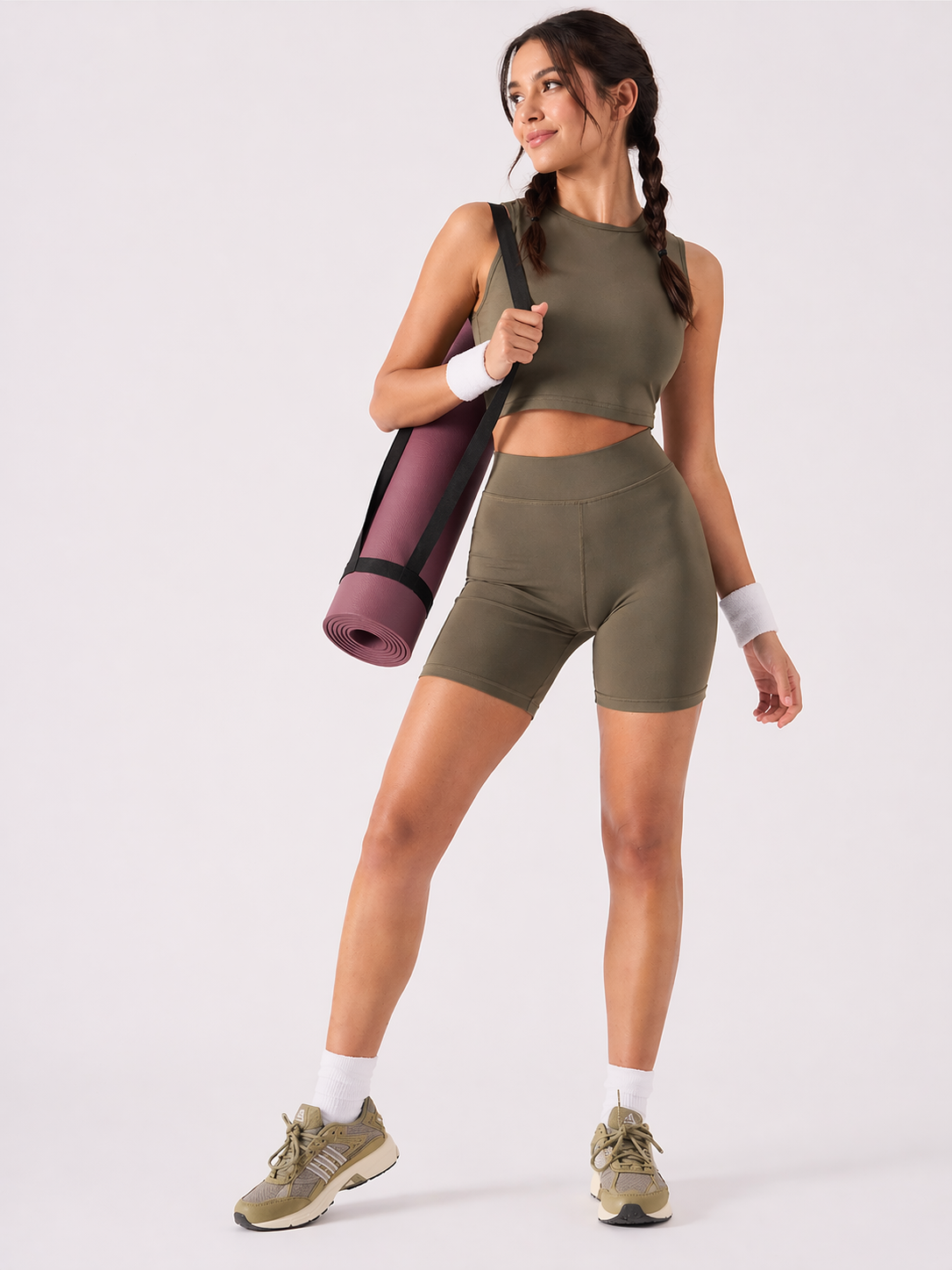 A woman wearing womens-khaki-high-waist-biker-shorts carrying a yoga mat, front view styled with a matching khaki crop top and olive trainers