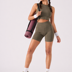 A woman wearing womens-khaki-high-waist-biker-shorts carrying a yoga mat, front view styled with a matching khaki crop top and olive trainers