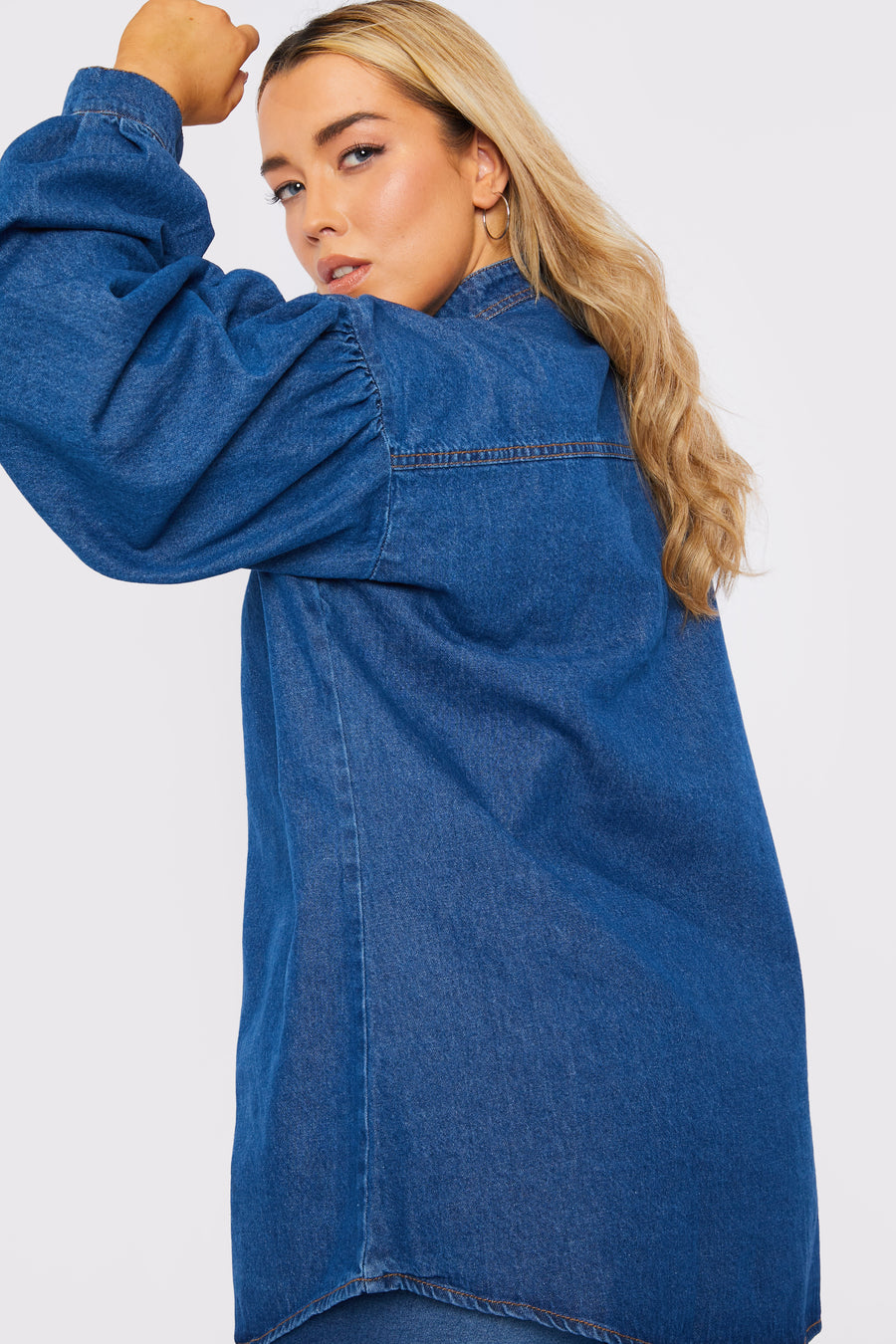 Side to back body shot of a standing plus size female model wearing a JMOJO Mid Blue Balloon Sleeve Denim Shirt