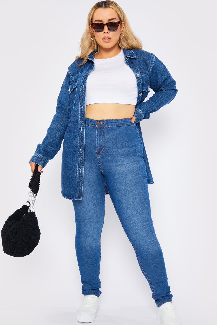 Full body styled shot of a standing plus size female model wearing JMOJO Mid Blue High Waisted Skinny Jeans holding a handbag and wearing sunglasses
