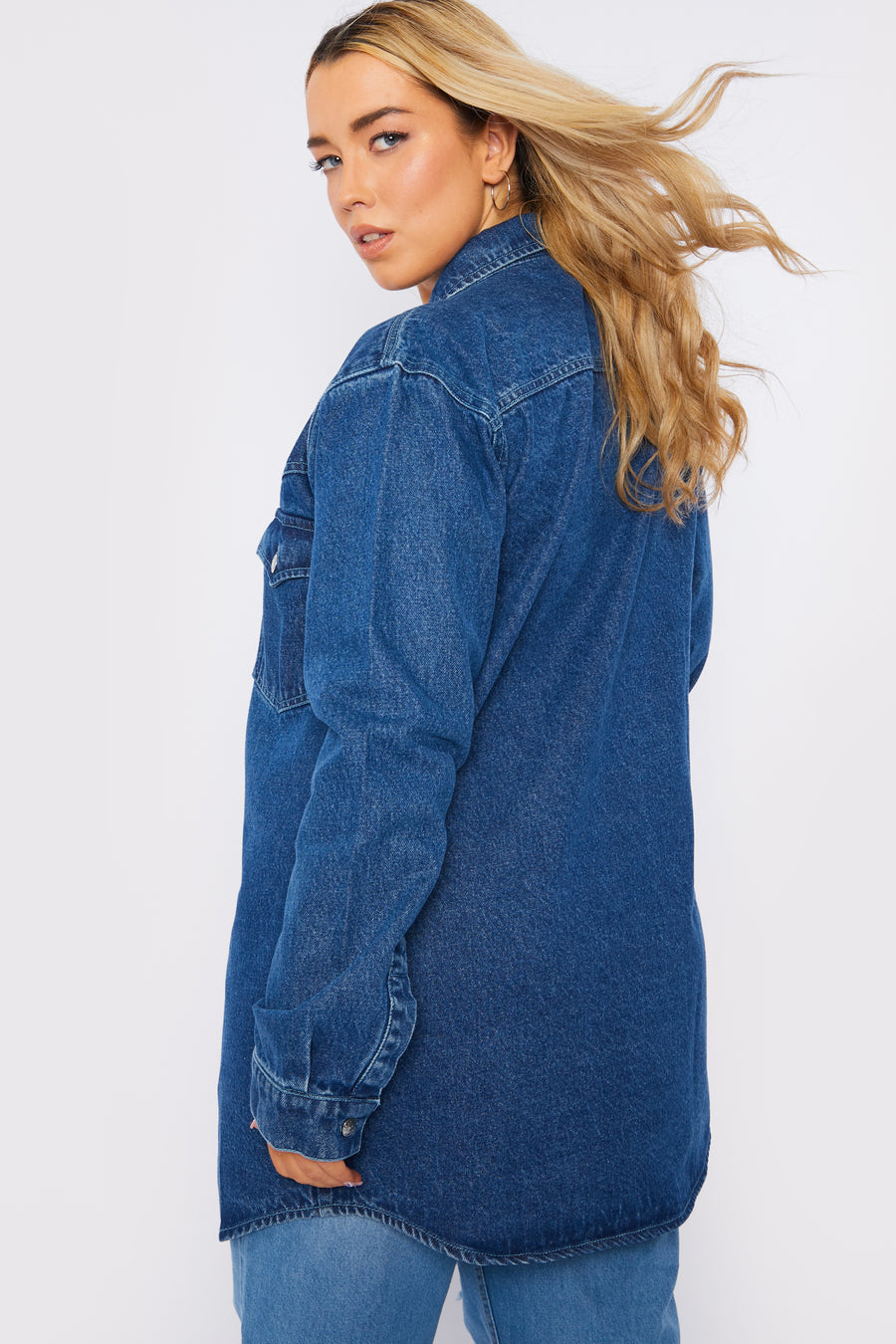 Body side to back shot of a standing plus size female model wearing a JMOJO Mid Blue Wash Long Sleeve Denim Shirt Dress