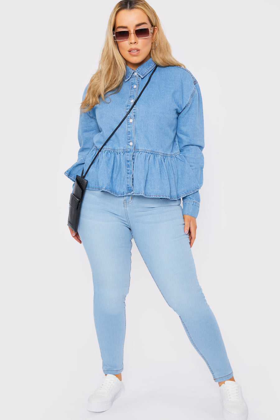 Full body style shot of a standing plus size female model wearing a JMOJO Light Blue Wash Peplum Denim Frill Shirt with a handbag over her shoulder and wearing sunglasses