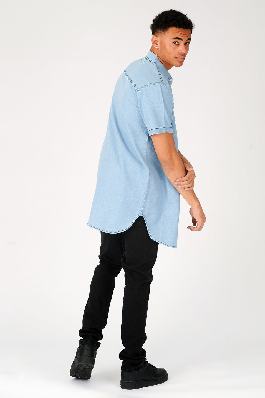 Body side shot of a standing male model wearing a JMOJO Light Blue Wash Slim Fit Longline Short Sleeve Denim Shirt