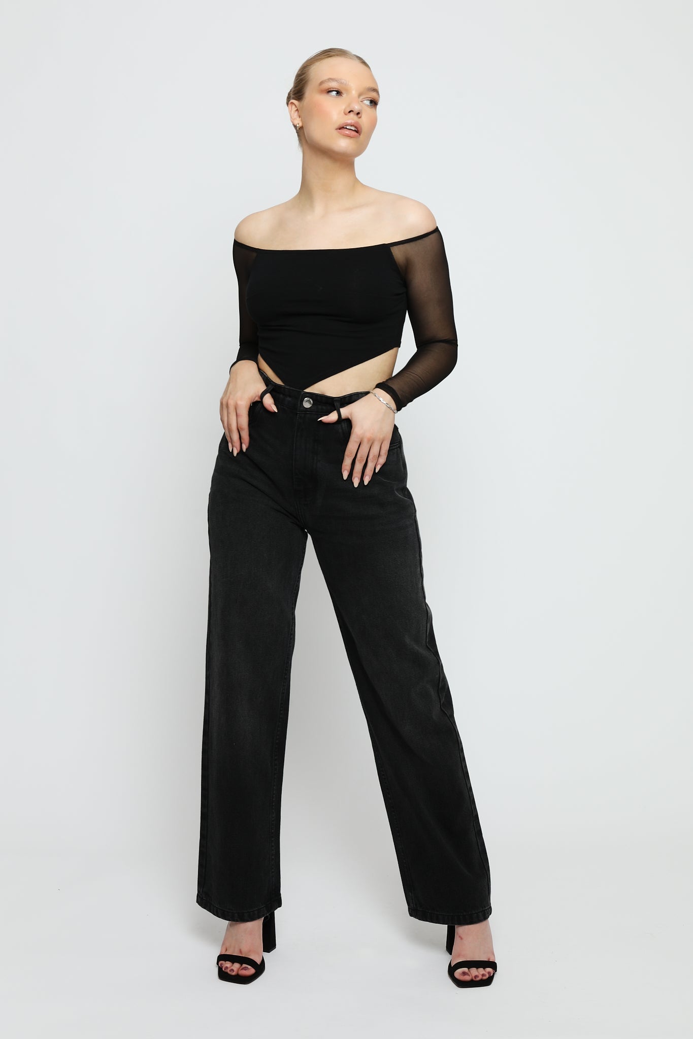 A woman posing in high-waisted black wide leg jeans with a black mesh top and heels.
