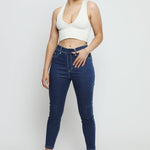 A woman wearing high-waisted dark blue denim jeggings with a white crop top and blue trainers.