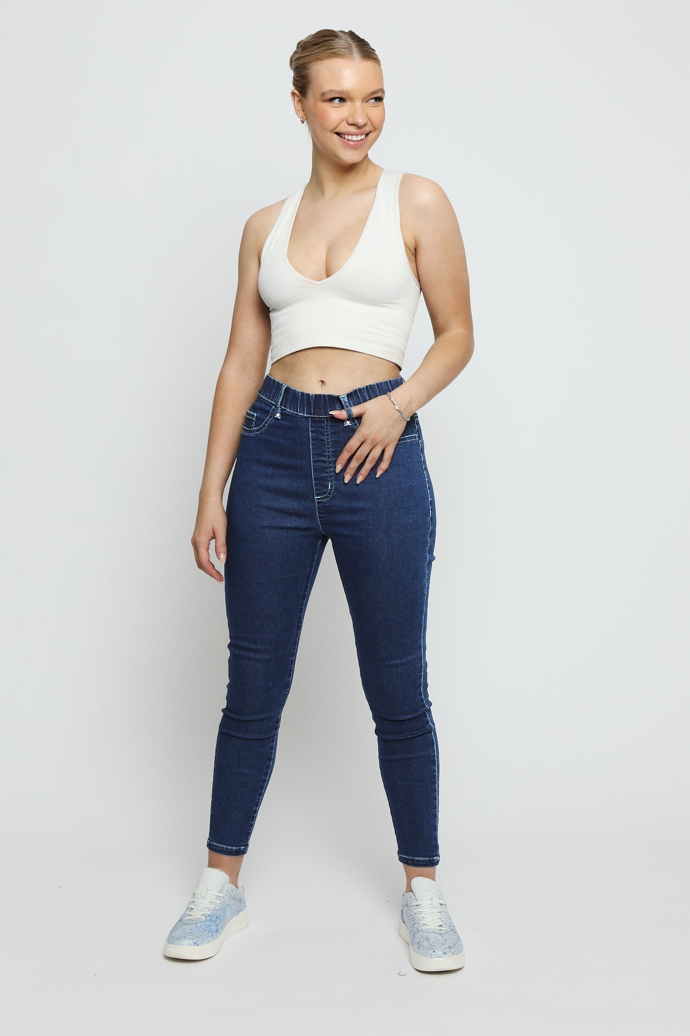 A woman wearing high-waisted dark blue denim jeggings with a white crop top and blue trainers.