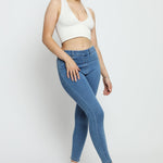 A woman wearing high-waisted mid blue denim jeggings with a white crop top and blue trainers.