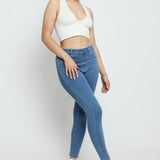 A woman wearing high-waisted mid blue denim jeggings with a white crop top and blue trainers.