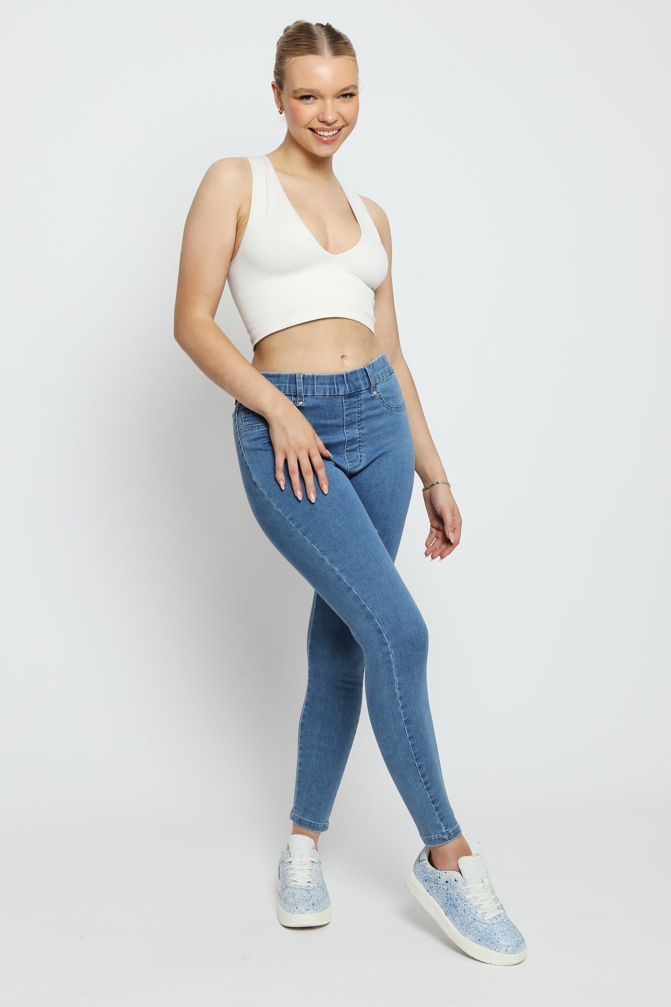 A woman wearing high-waisted mid blue denim jeggings with a white crop top and blue trainers.